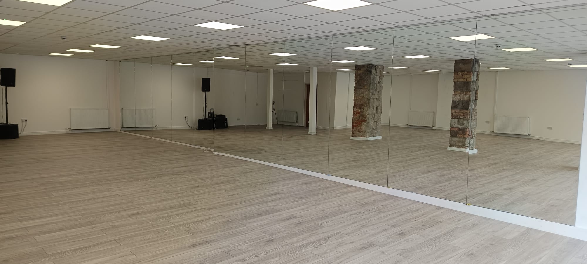 The Peters Hill room empty and lit, its mirrored wall folding the pale wood floor and brick columns back on themselves.