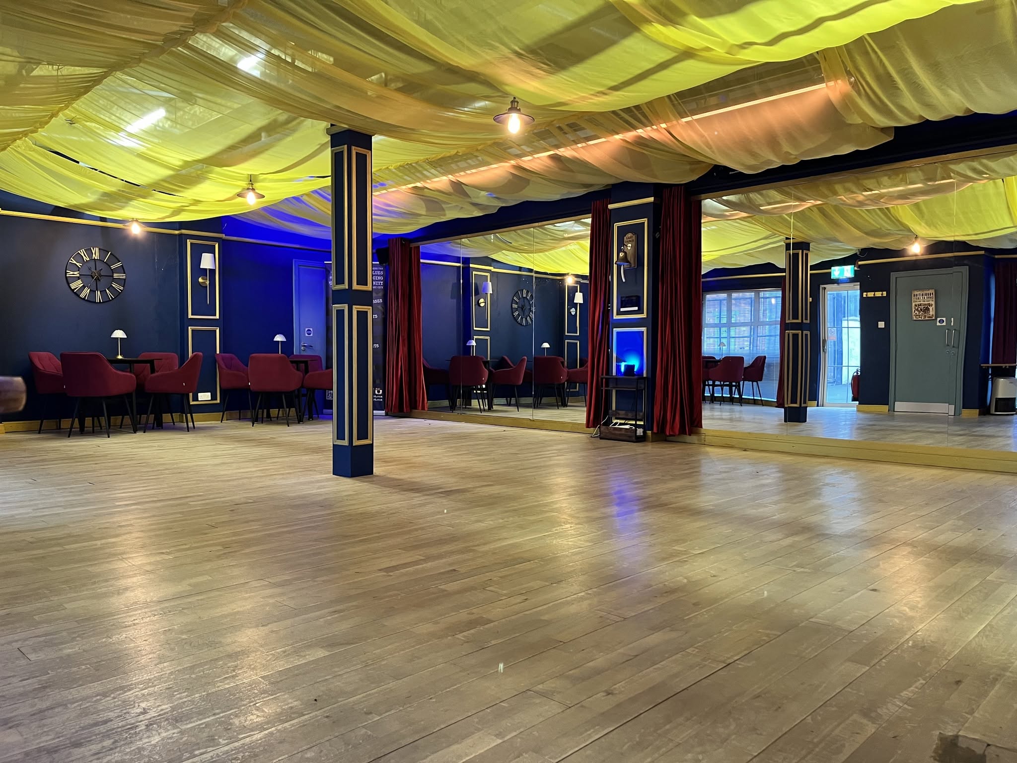 The Social Space ballroom: a wide sprung wood floor, navy and gold panelled walls, red velvet chairs at tables down one side and cream fabric draped across the whole ceiling.