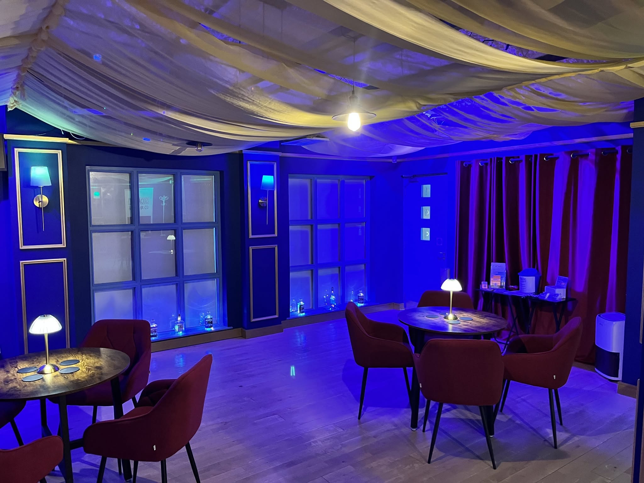 A lounge at Social Space washed in deep blue light: red armchairs around low tables, brass lamps lit, cream fabric draped overhead.