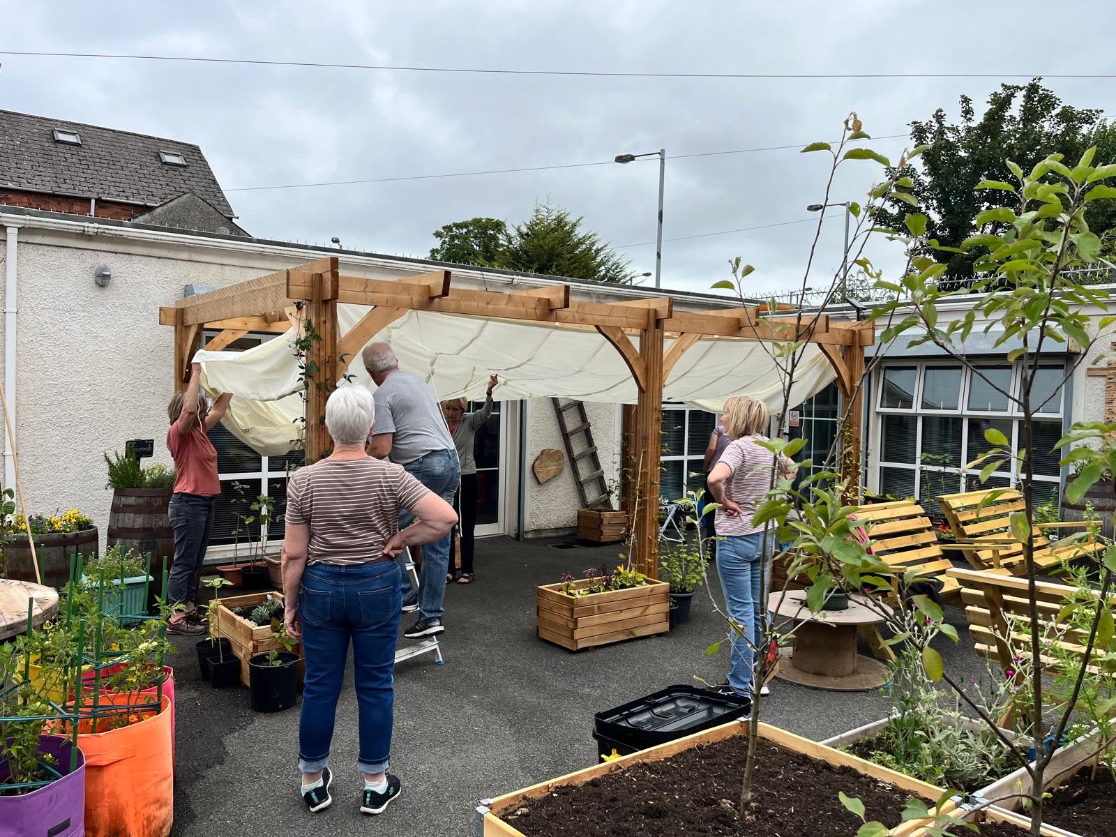 Half a dozen people in the Social Space yard raising a cream canvas sail over a new timber pergola, surrounded by raised beds, planters and fruit trees.